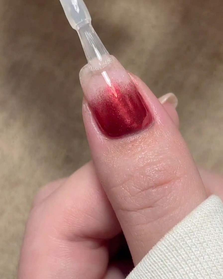 A brush applying a clear sealing coat of base gel over a red gradient nail design.