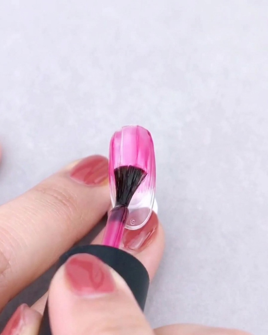A fan brush gently blending a pink gel color downward on a clear nail to create a gradient effect.