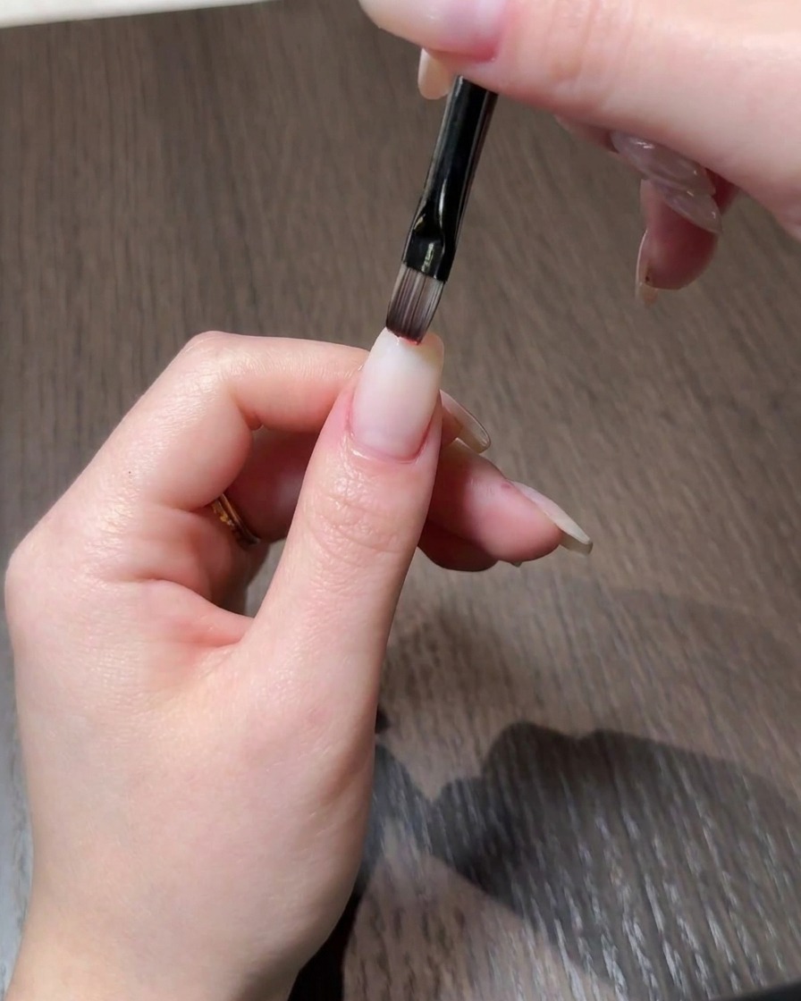 A close-up of a hand applying red gel polish to the tip of a natural-looking nail using a fine brush.