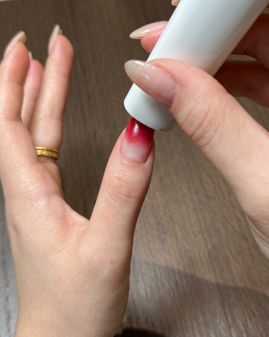 A thumb positioned directly under a portable white UV and LED nail lamp to cure the red ombre gel.
