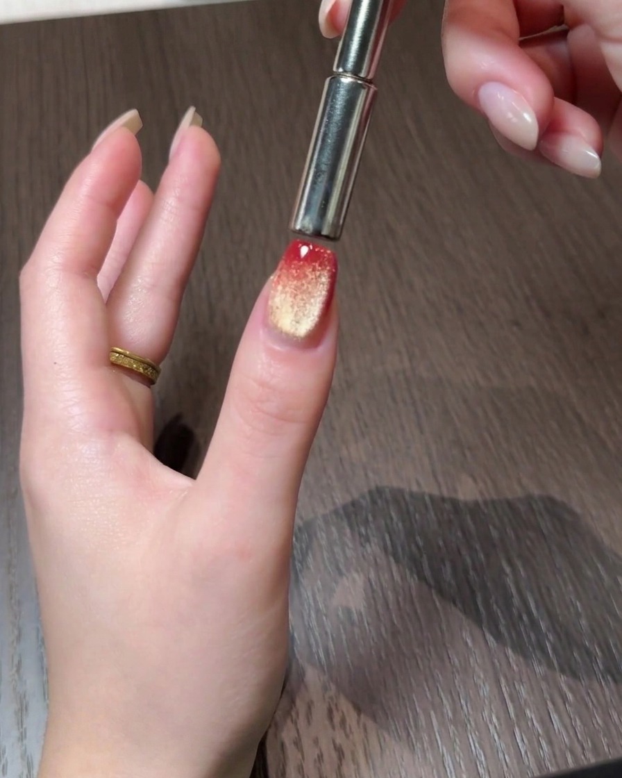 A magnetic tool pushing cat eye particles toward the back edge of a red and gold gradient nail.