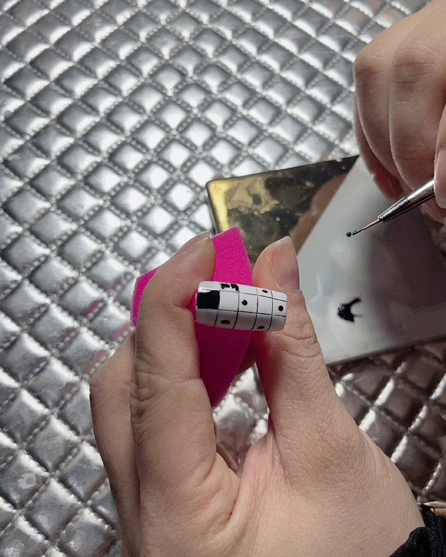 A close-up of a nail artist using a fine liner brush to straighten the edge of a black square on a checkerboard nail design.
