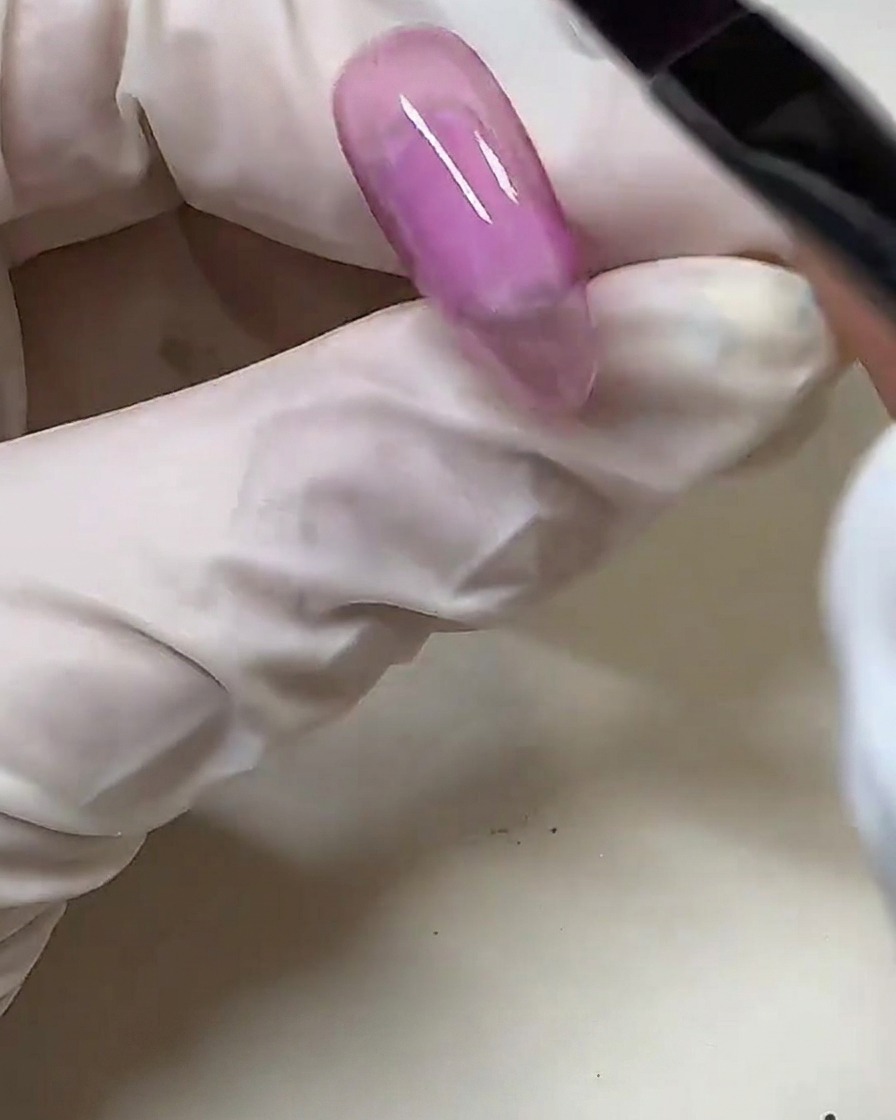 A gloved hand using a gel brush to apply semi-transparent pink gel polish over a clear nail tip.