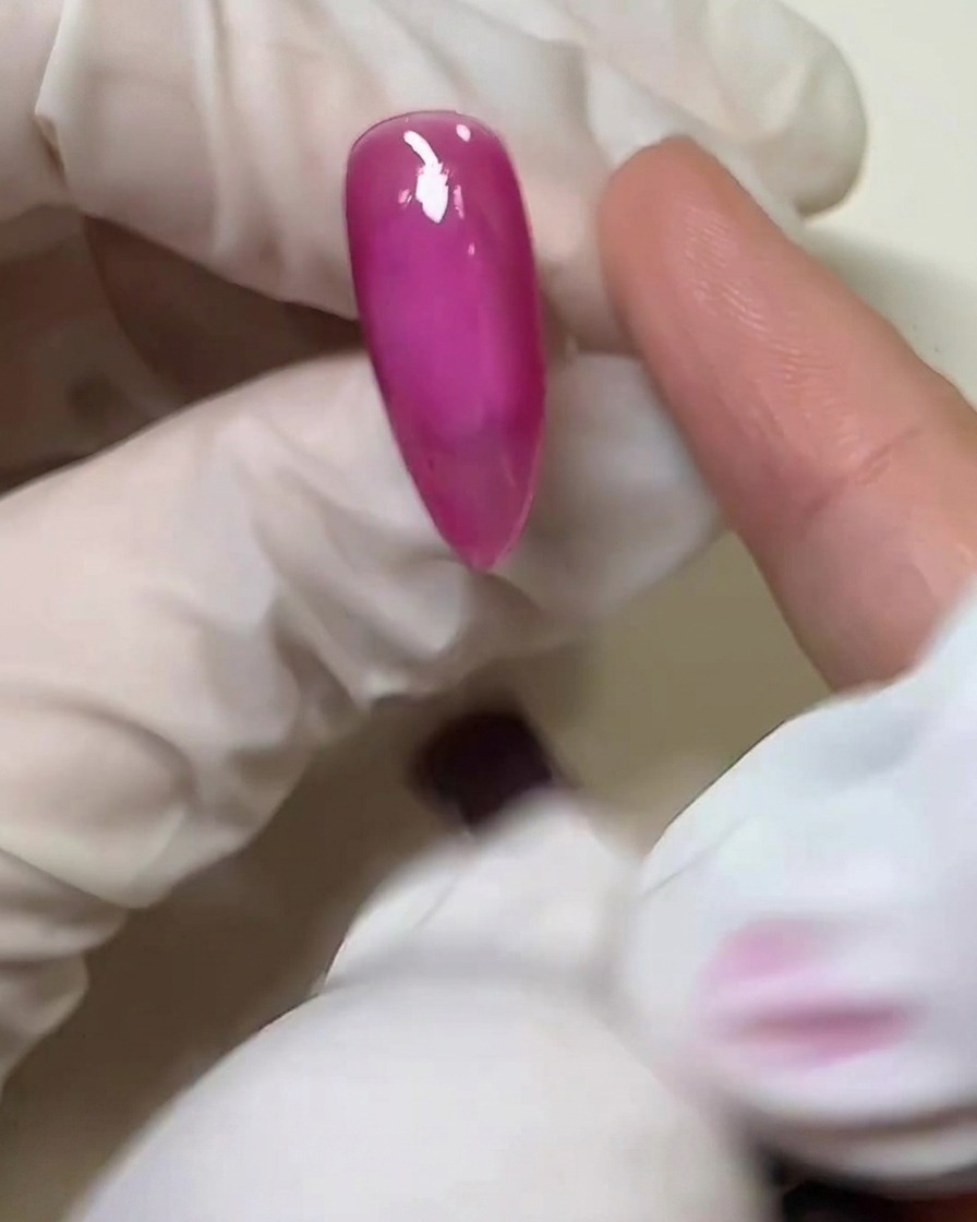 A close-up of a gloved finger applying a thin layer of clear solid glue over the cured pink nail surface.