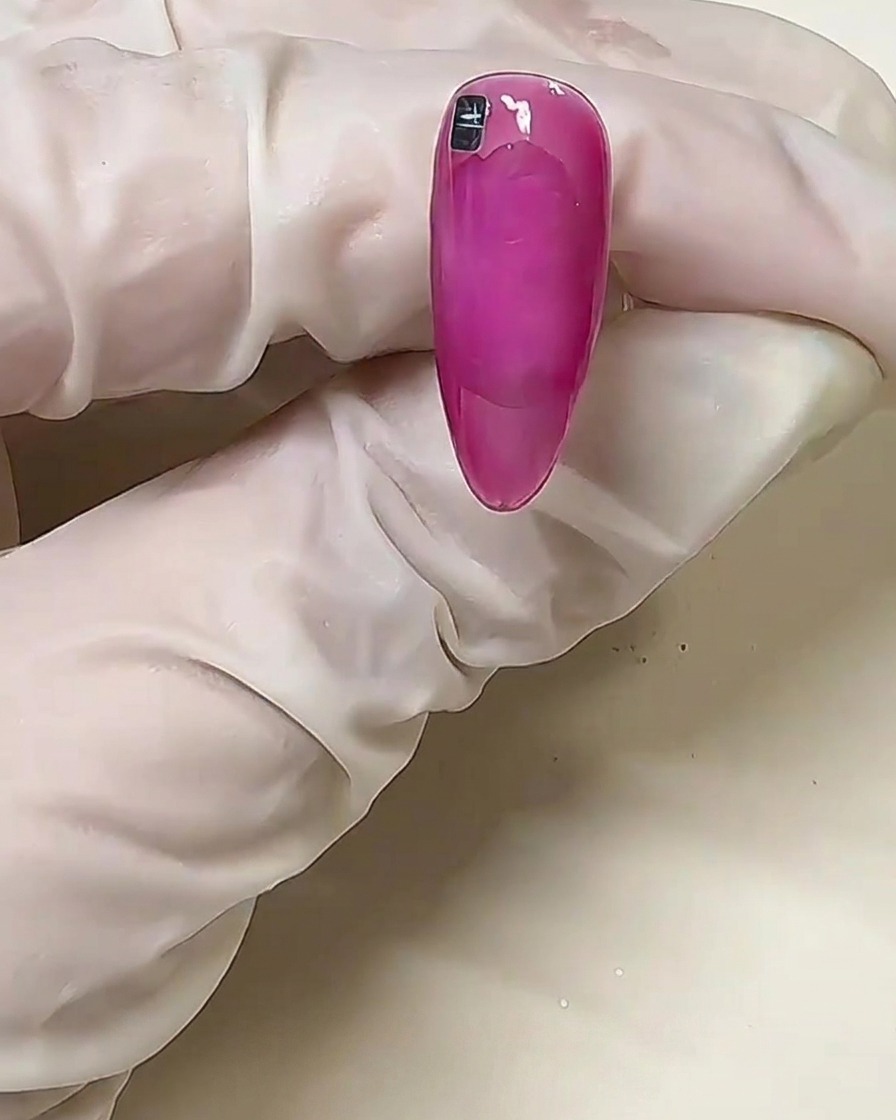 A close-up of a pink nail with a single silver metallic square placed near the cuticle line on uncured solid glue.