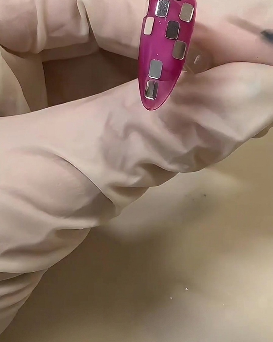 A gloved hand adjusting small silver square foil pieces on a translucent pink nail to create a checkerboard pattern.