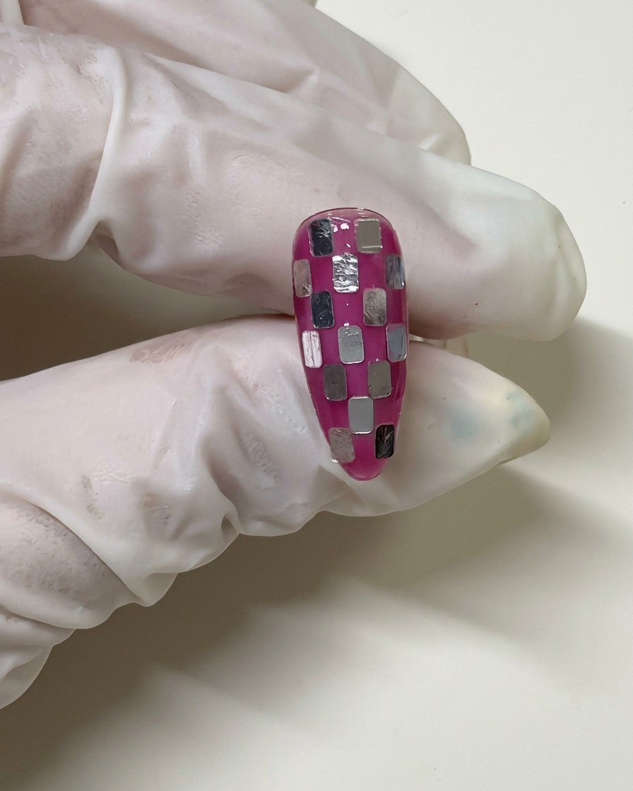 A gloved hand holding a nail with a checkerboard pattern that has been coated in a sheer pink gel layer to blend the silver squares.