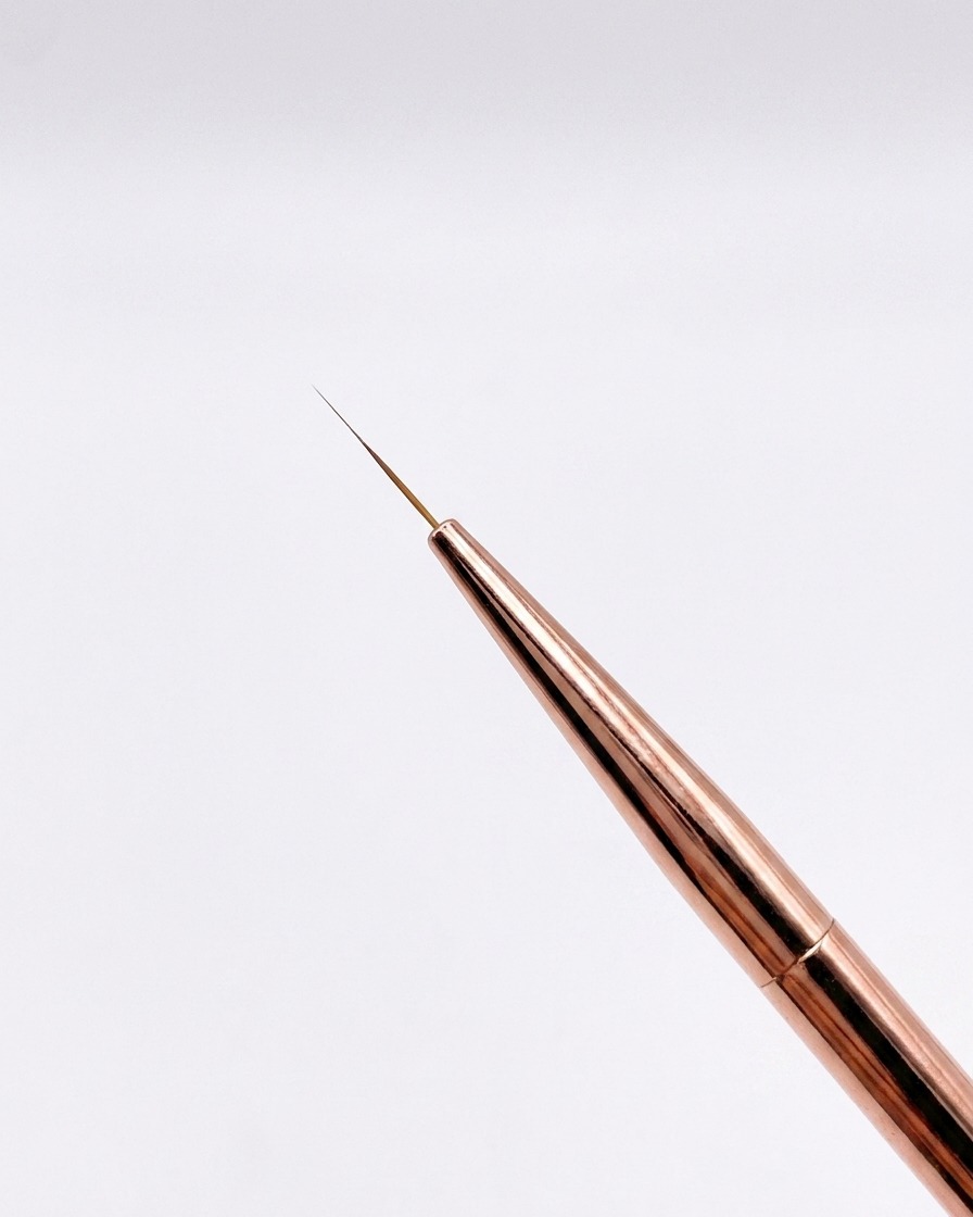 A close-up view of a fine 12mm rose-gold liner brush with a sharply pointed tip against a neutral background.
