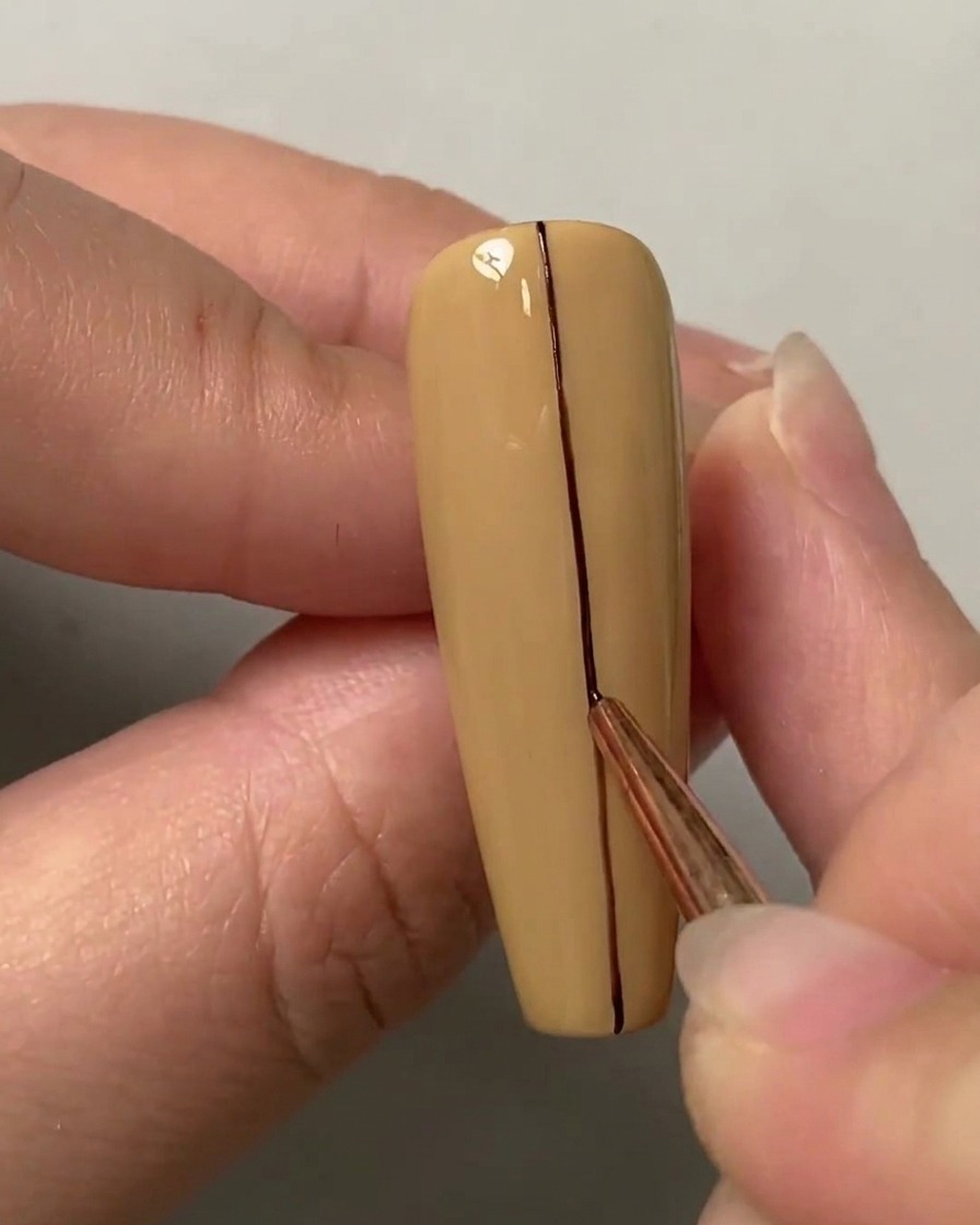 A liner brush tracing a straight dark line down the vertical center of a nude-colored nail.