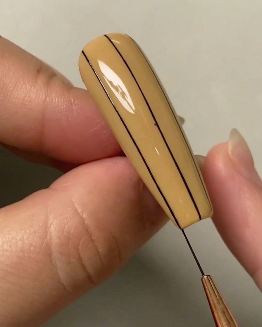A close-up of a liner brush drawing a third vertical black line on the right side of a nude-colored nail.