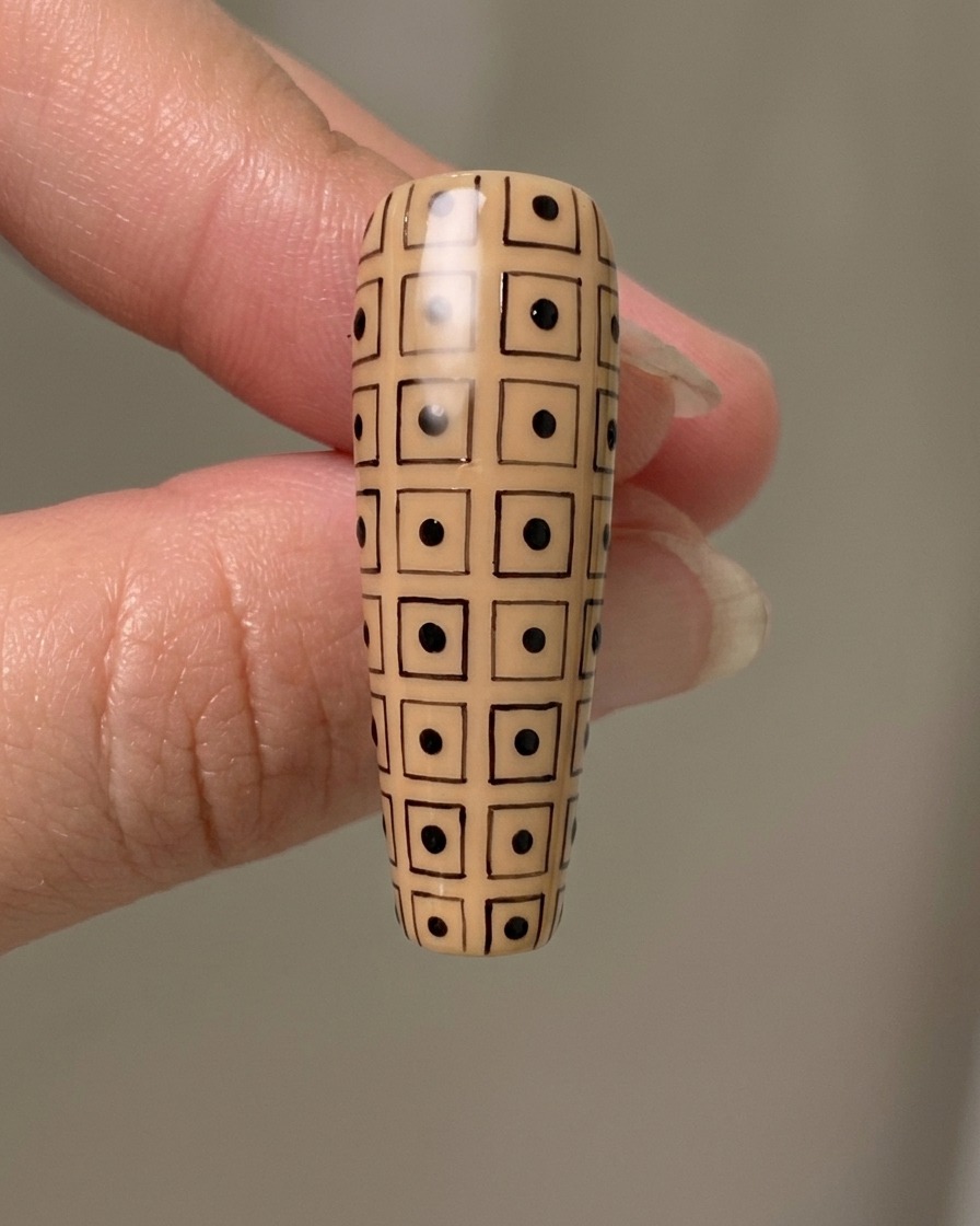 A long nude nail with a hand-drawn black grid, featuring alternating black dots inside the squares.