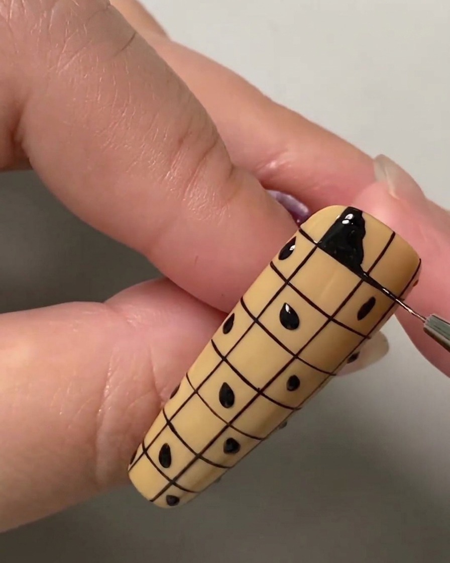 A close-up of a liner brush filling in the top left square of a grid on a nude nail with black gel polish.