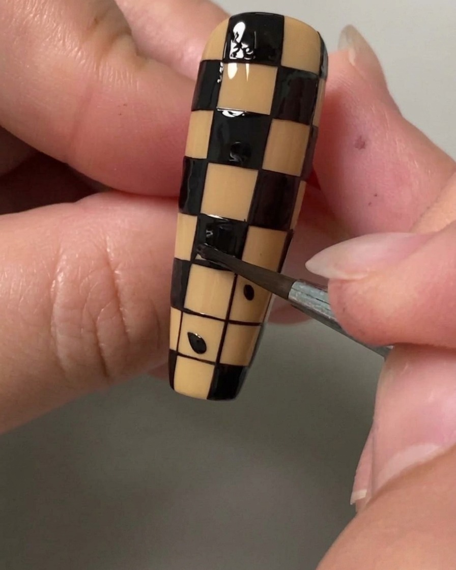 A close-up of a fine detail brush filling a small square with black polish to create a checkerboard pattern on a nail.