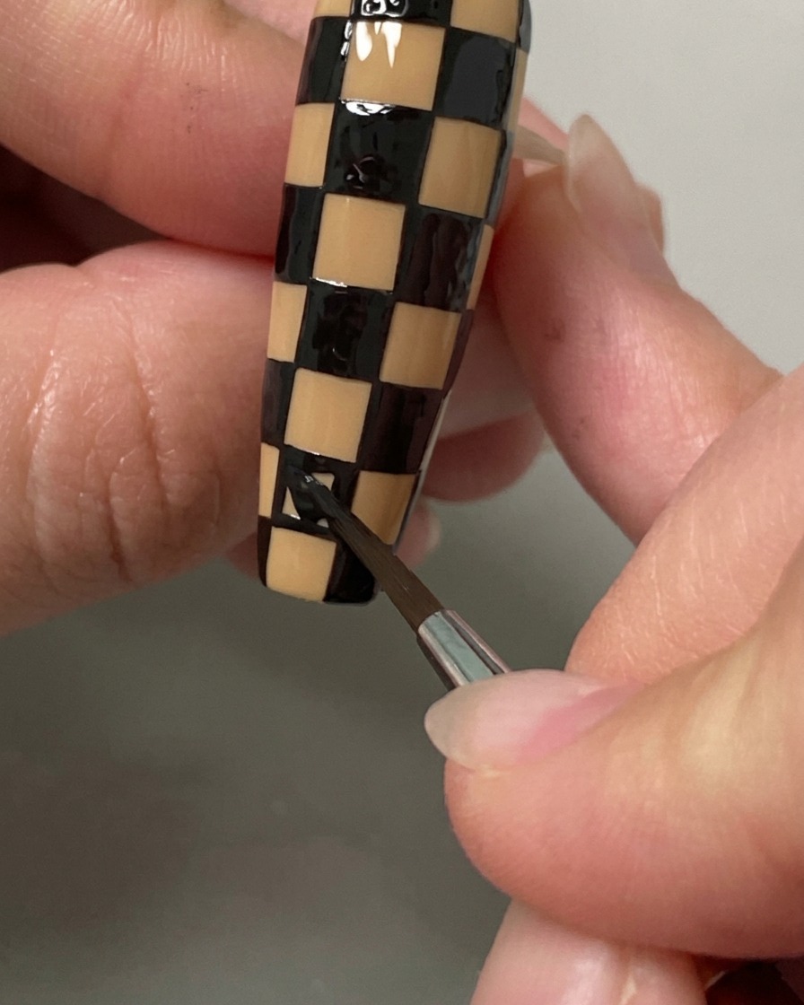 A liner brush filling in the final squares of a black and nude checkerboard pattern on a long nail.