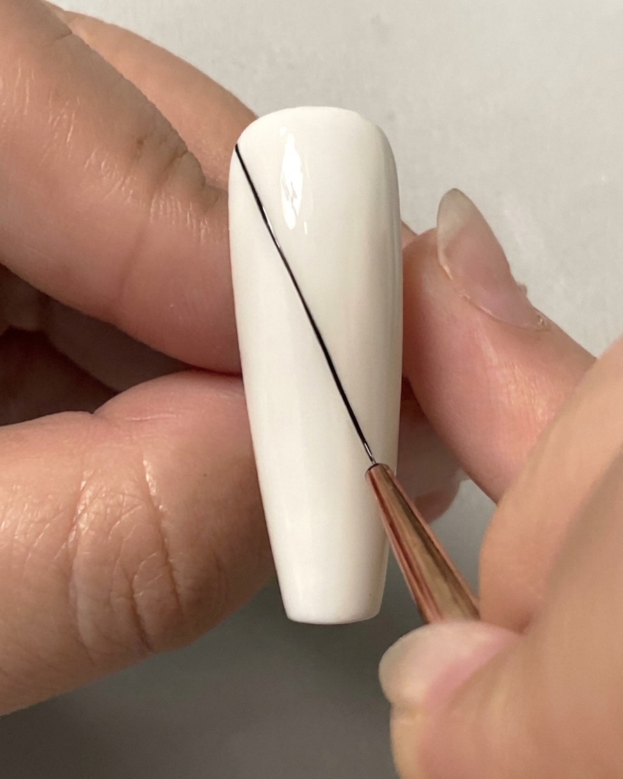 A close-up of a fine liner brush drawing a long diagonal black line across a white nail.