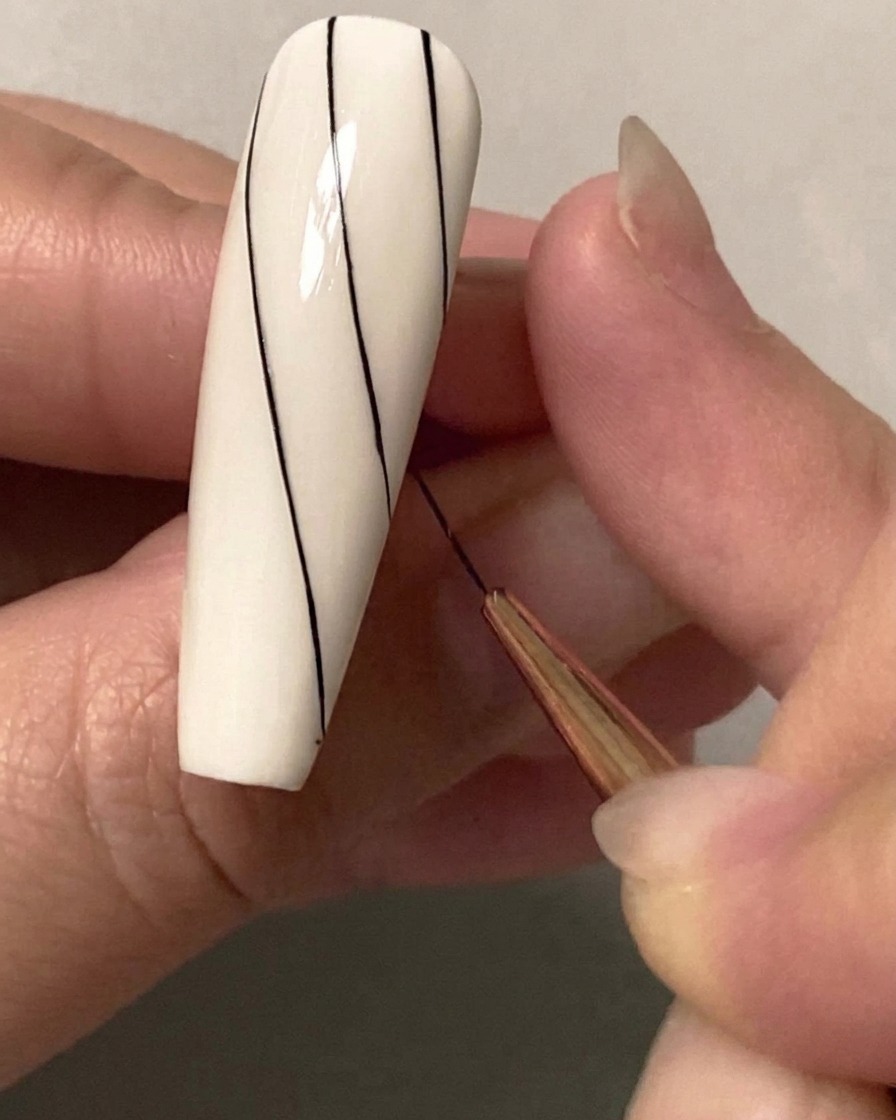 A close-up of a fine nail art brush drawing a second parallel diagonal black line on the upper half of a white nail.