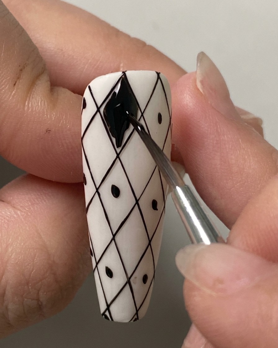 A fine liner brush carefully filling in the uppermost diamond shape with black gel polish on a pre-drawn white grid nail.