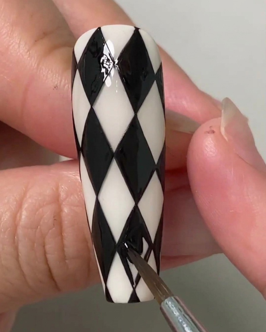 A nail artist using a fine brush to fill the last remaining diamond shapes on a black and white argyle patterned nail.