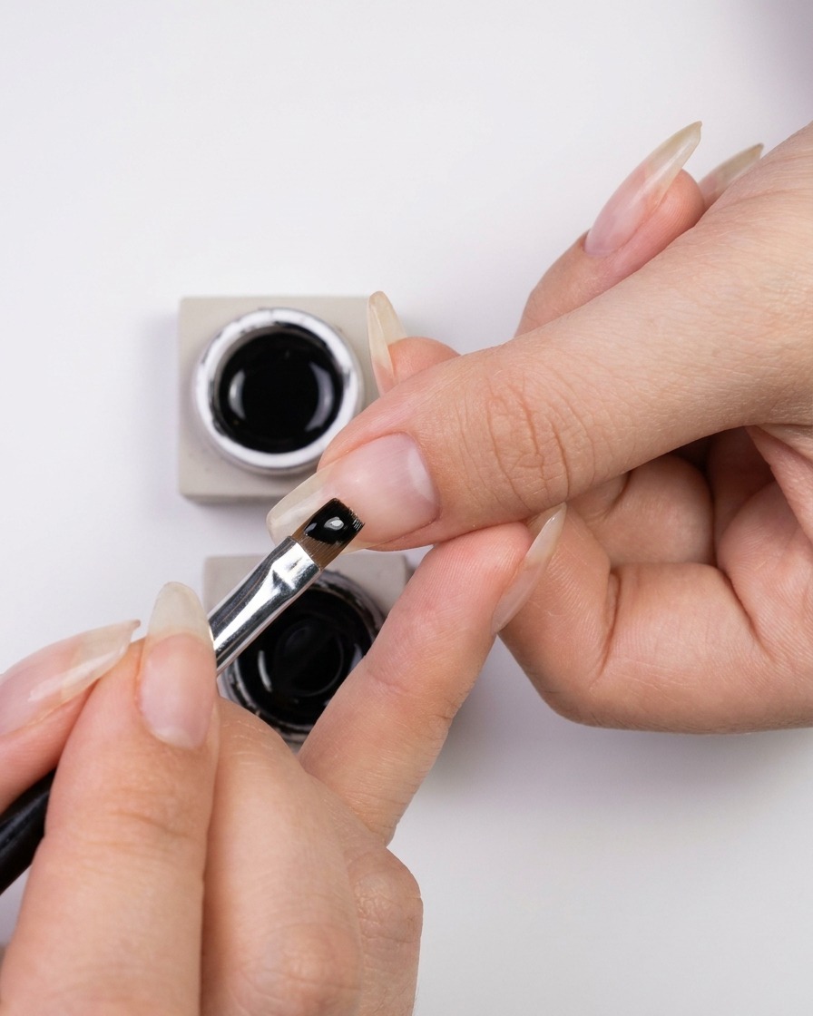 A hand dipping a fine nail art liner brush into a small jar of glossy black gel polish.