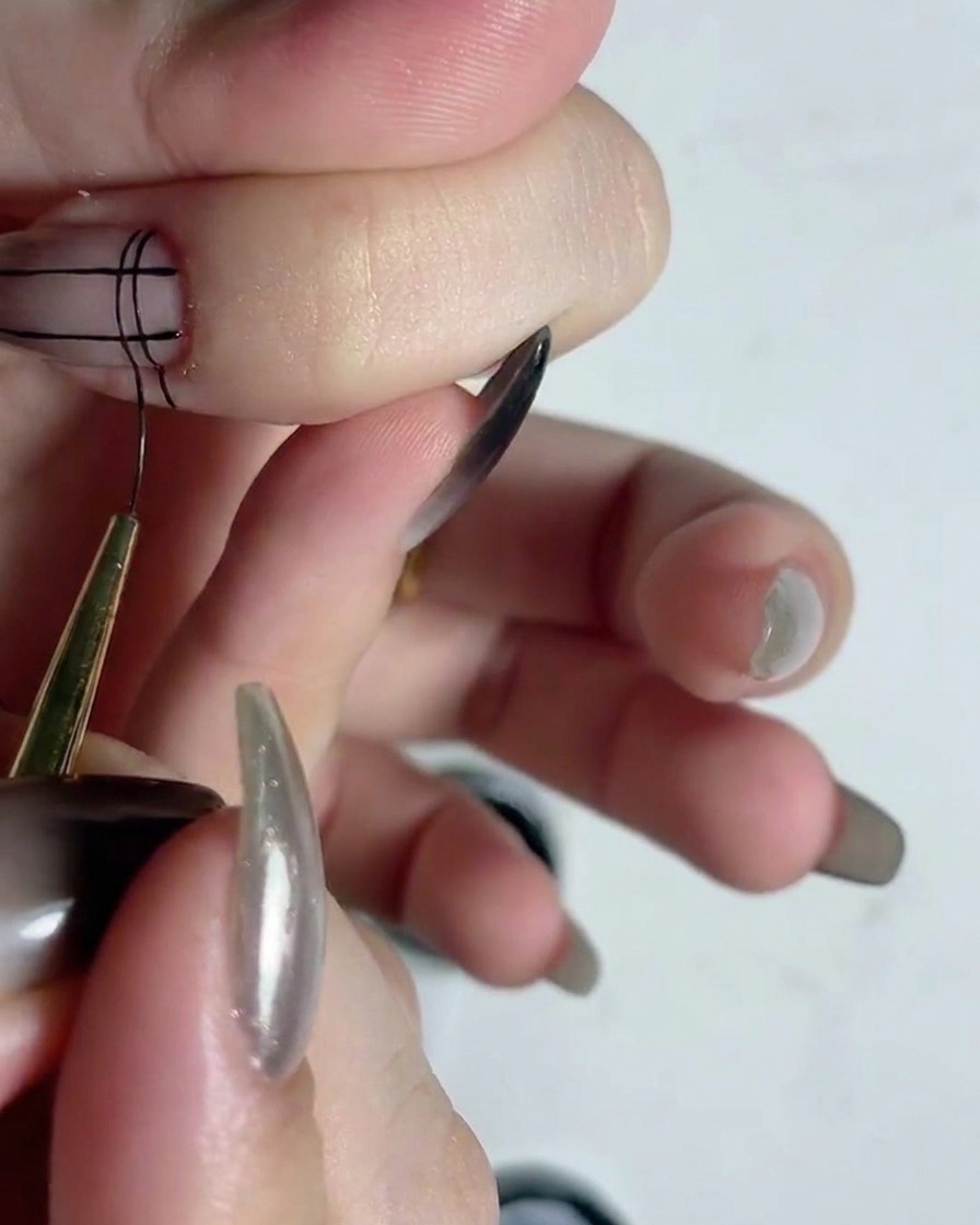 A fine liner brush drawing horizontal black lines over vertical lines to create a grid pattern on a fingernail.