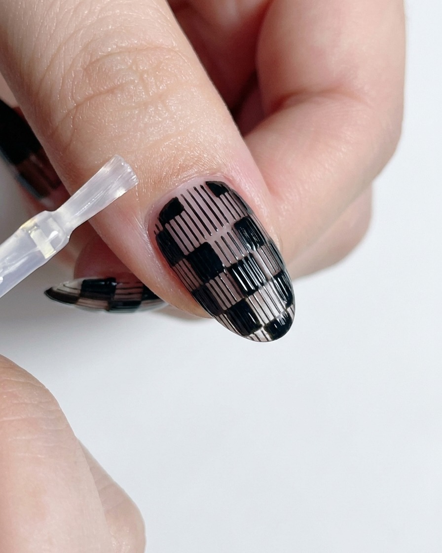 A top coat brush applying clear gloss polish over a finished black and white checkered nail.