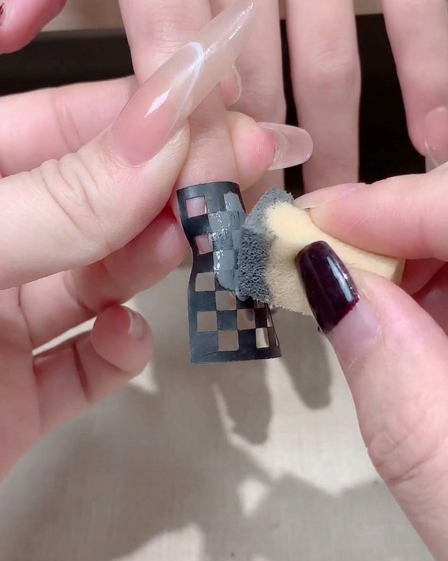 A close-up of a manicurist adjusting a black and white checkered nail sticker on a long square nail.