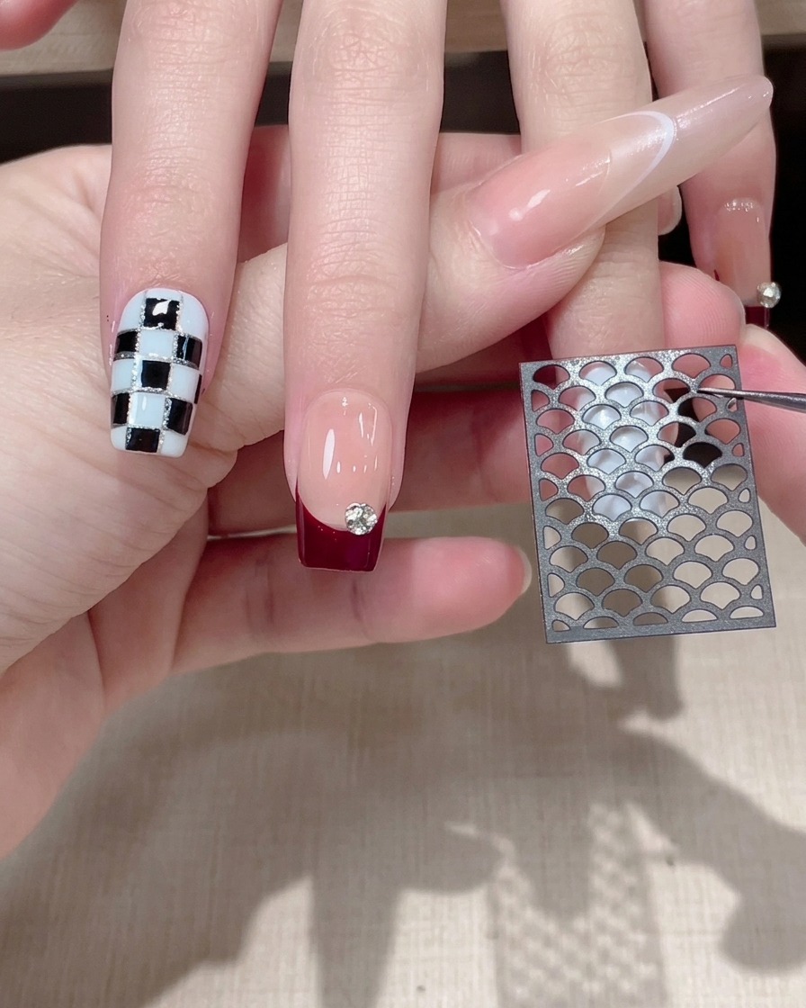 Tweezers holding a hollow black mesh sticker over a nude and burgundy French-tipped nail.