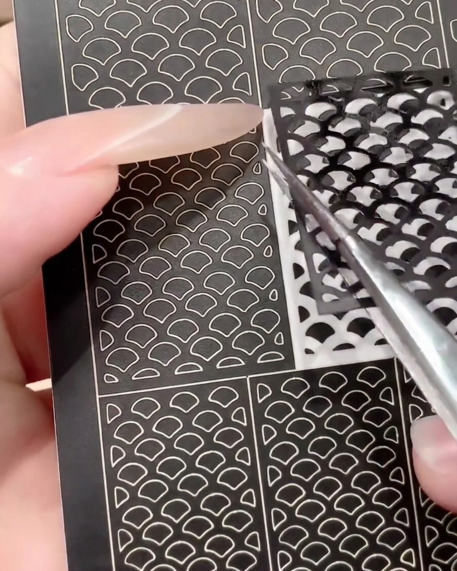 A pair of tweezers peeling a used black nail art sticker sheet to show reusability.