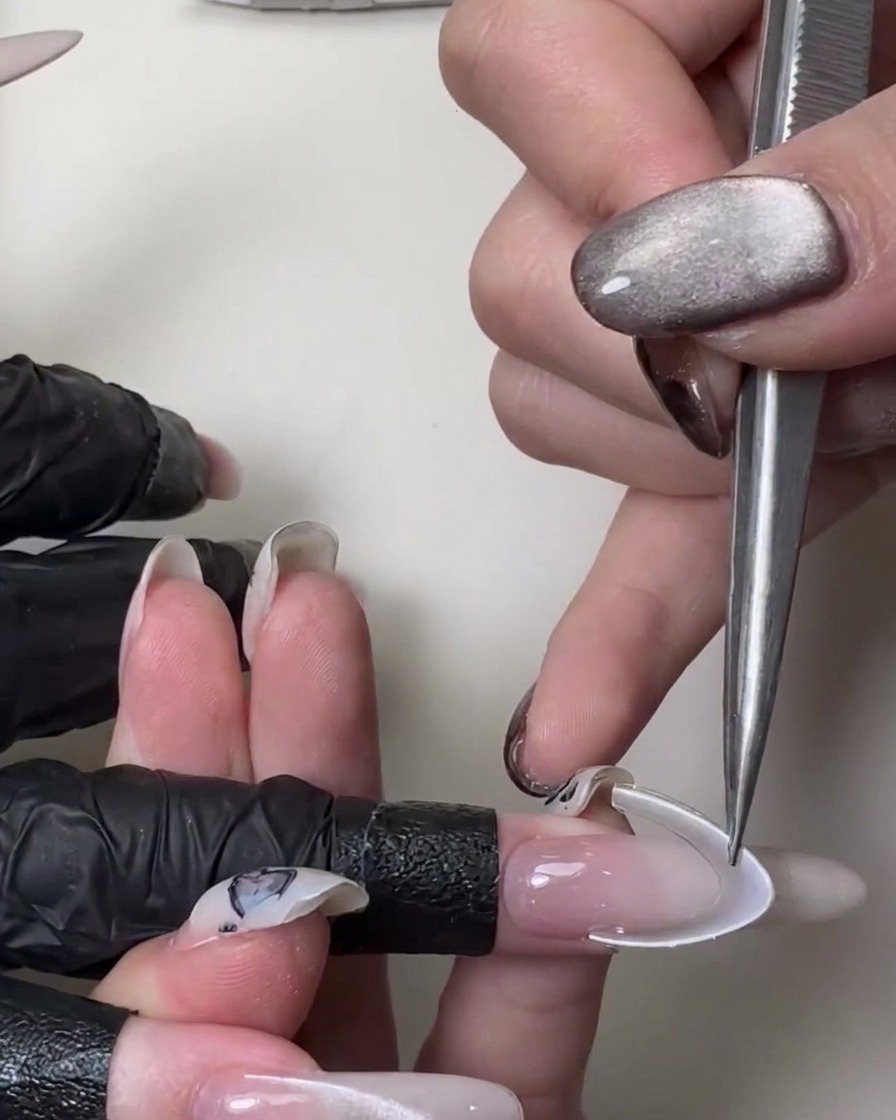 A close-up of a hand using metal tweezers to apply a white curved French smile line sticker onto a long, sheer pink stiletto nail.