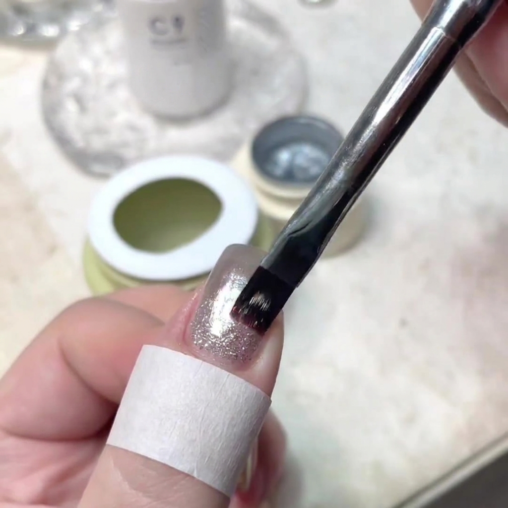 A nail brush precisely applying a thin stroke of shimmering gel to the center of a manicured nail.