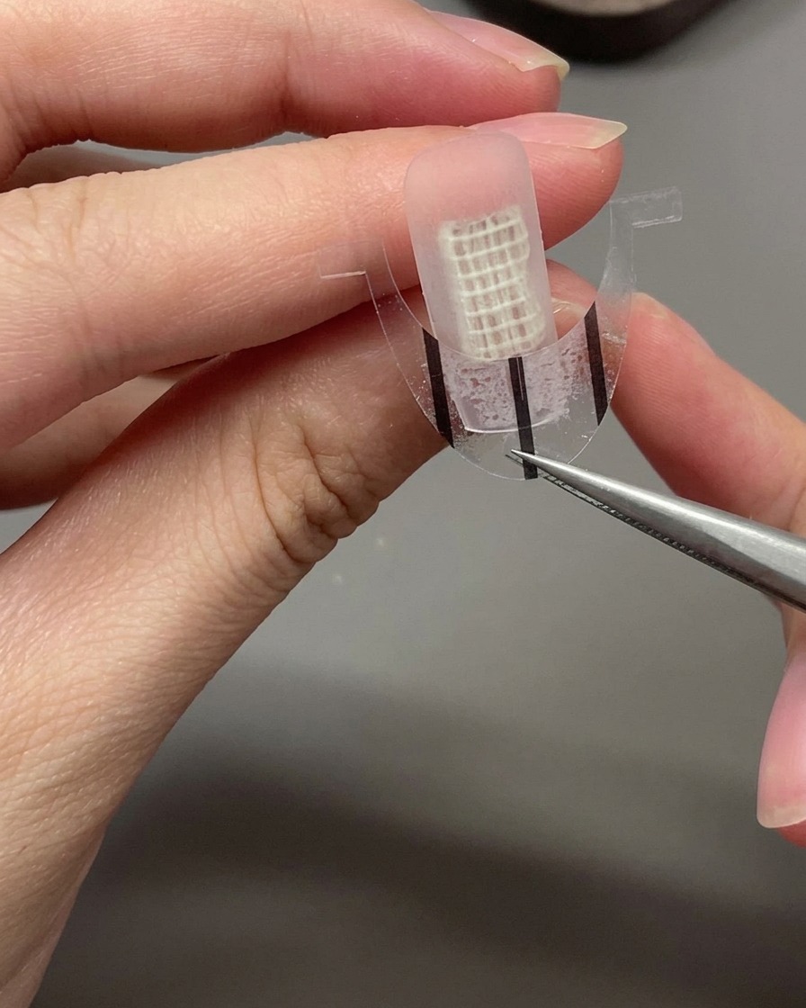 A pair of tweezers carefully placing a curved French guide sticker across the center of a bare nail.