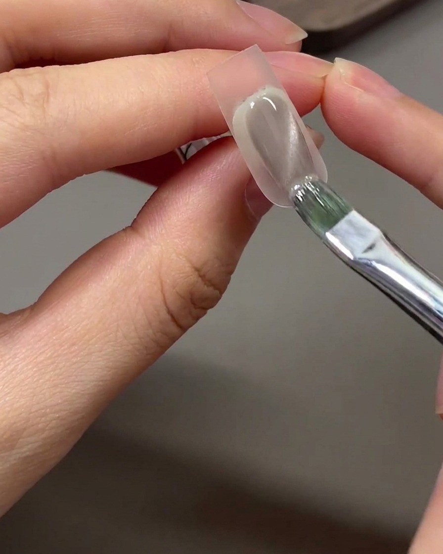 A brush applying grey-nude cat eye gel polish over the sponge-dabbed tip area of a nail.