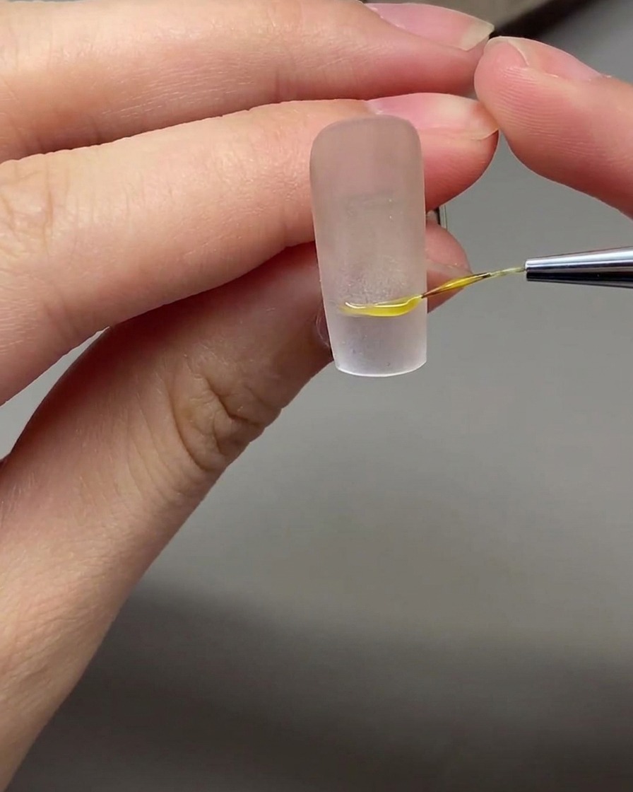 A fine nail art brush drawing a precise white French smile line on a nude base nail.