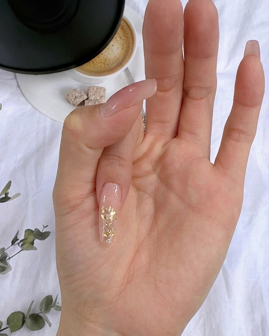 A close-up of gold foil flakes being pressed onto the wet glitter layer of a fingernail.