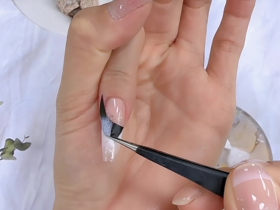 A pair of tweezers peeling off a black French guide sticker from a nail to reveal the gradient design underneath.