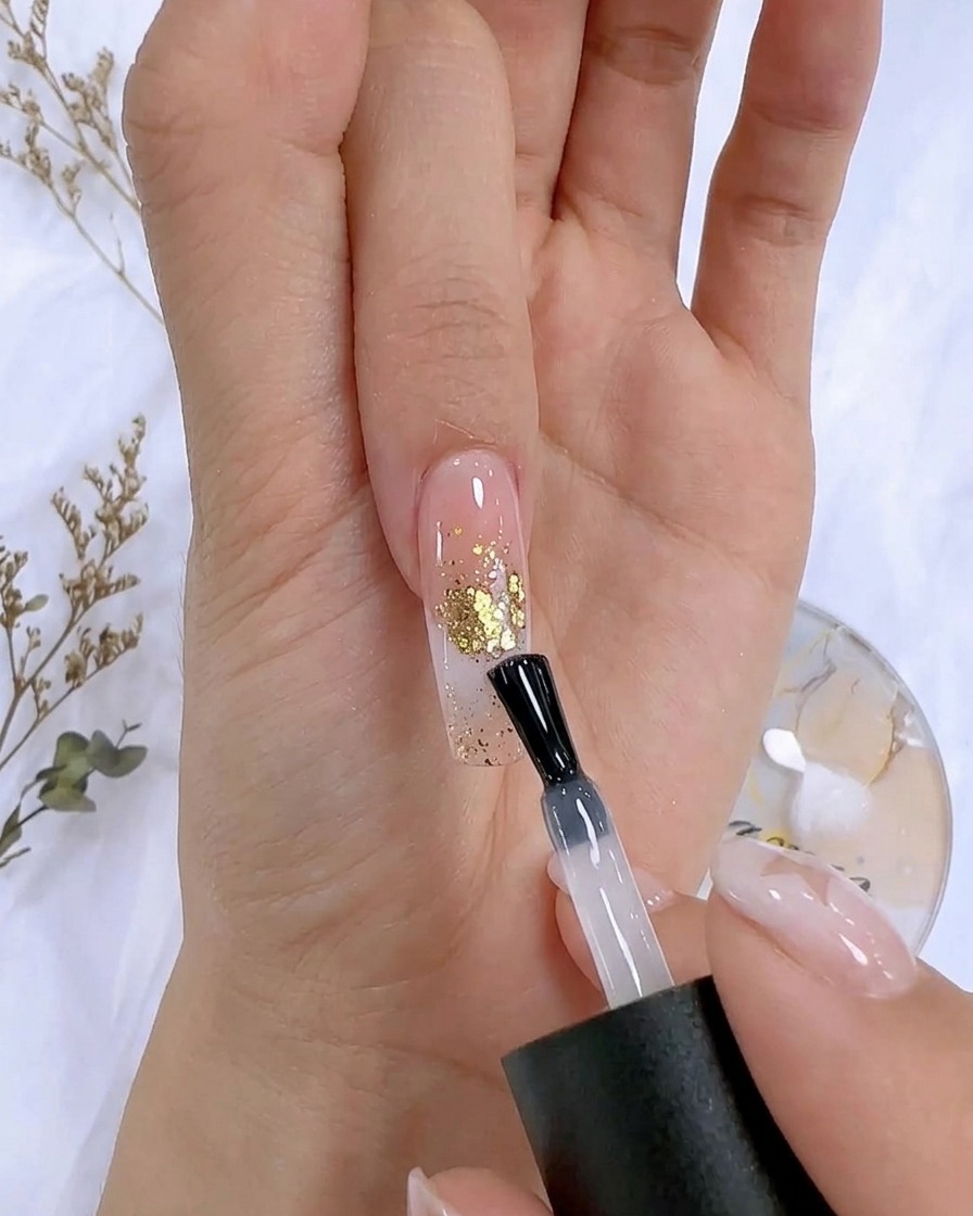 A nail polish brush applying a clear top coat over a decorated nail with gold foil accents.