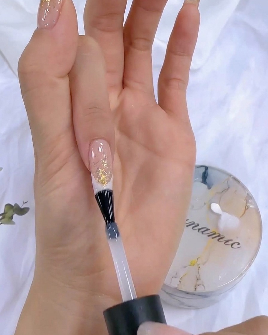 A hand holding a top coat bottle near a UV/LED nail lamp during the final curing stage.