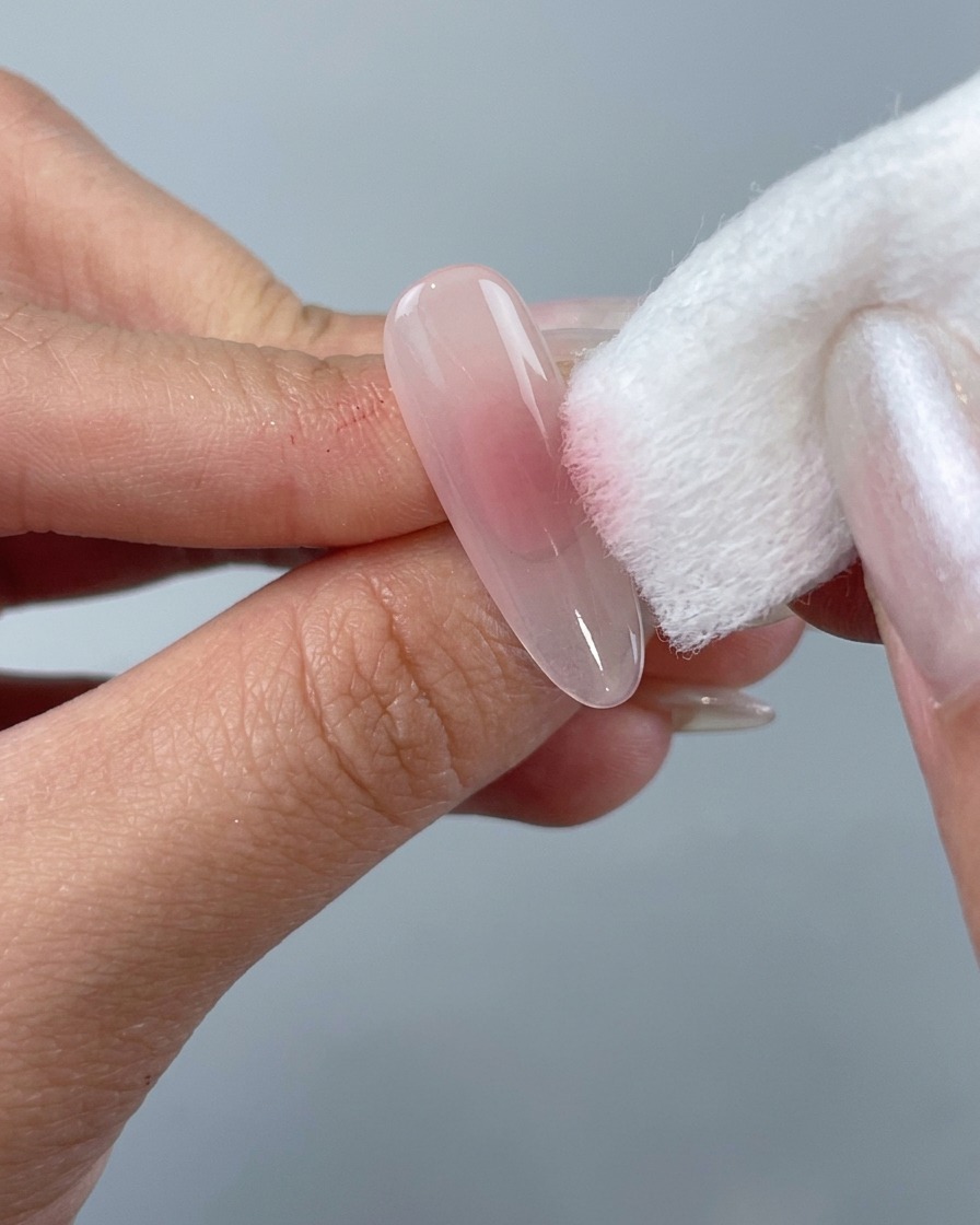 A hand using a white sponge block to dab gentle pink gel polish onto the center of a clear almond nail tip.