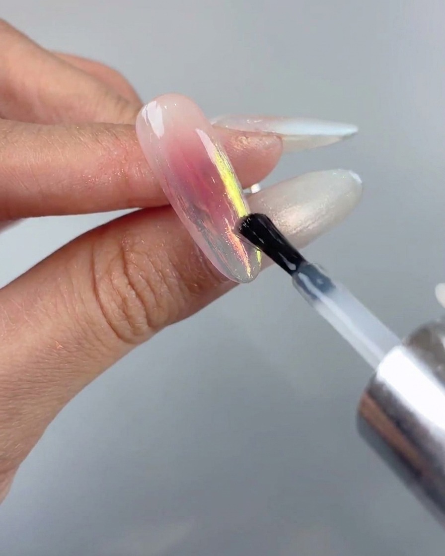 A nail brush applying a final sealing layer of smooth top coat over an aurora chrome nail design.