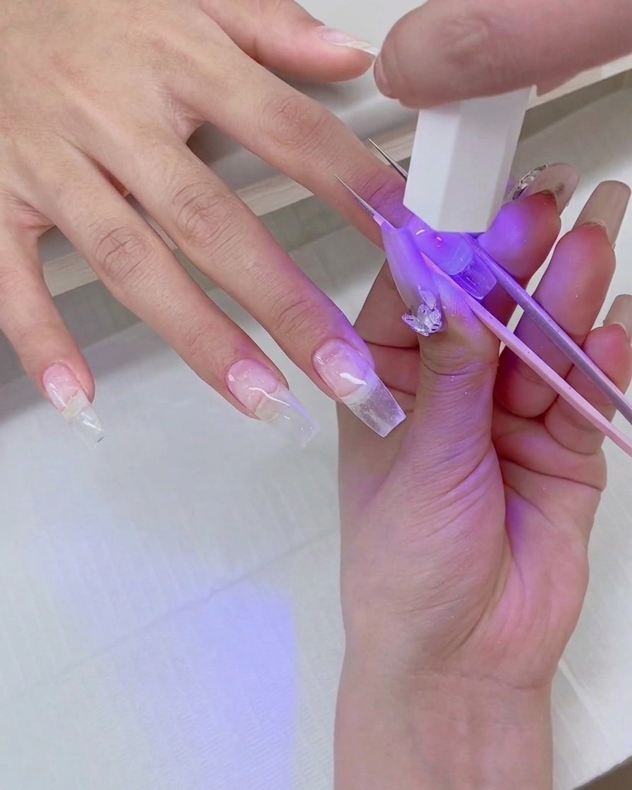 A close-up of a hand holding a mini UV flash-cure lamp over a clear nail extension while tweezers stabilize the tip.