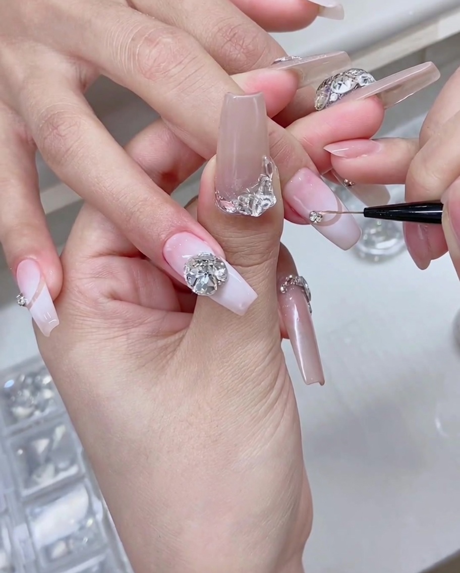 A fine detail brush and tweezers placing sparkling silver rhinestones onto the base of a manicured nail.