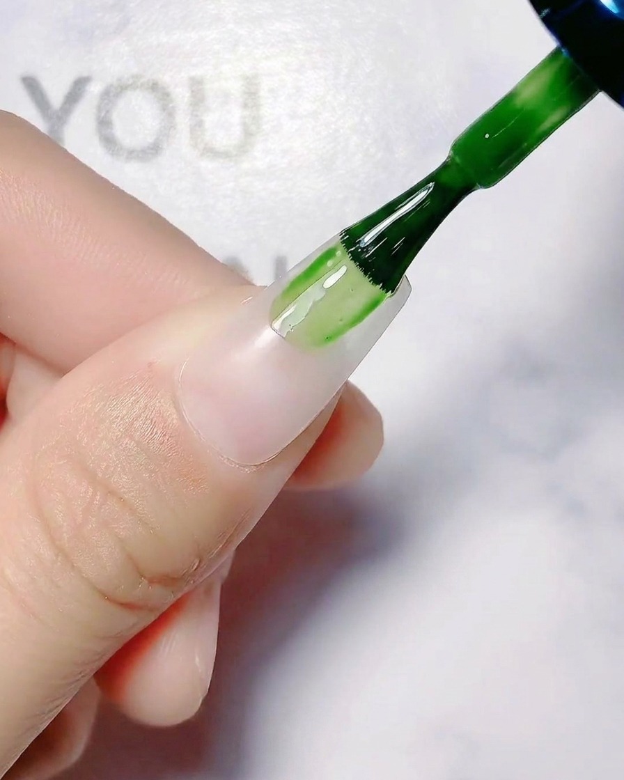 A nail brush applying translucent white base coat onto a bare fingernail.