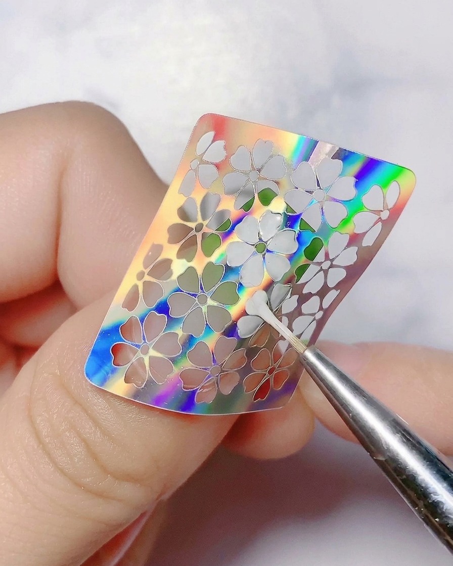A fine tool peeling away a holographic flower sticker from the nail.