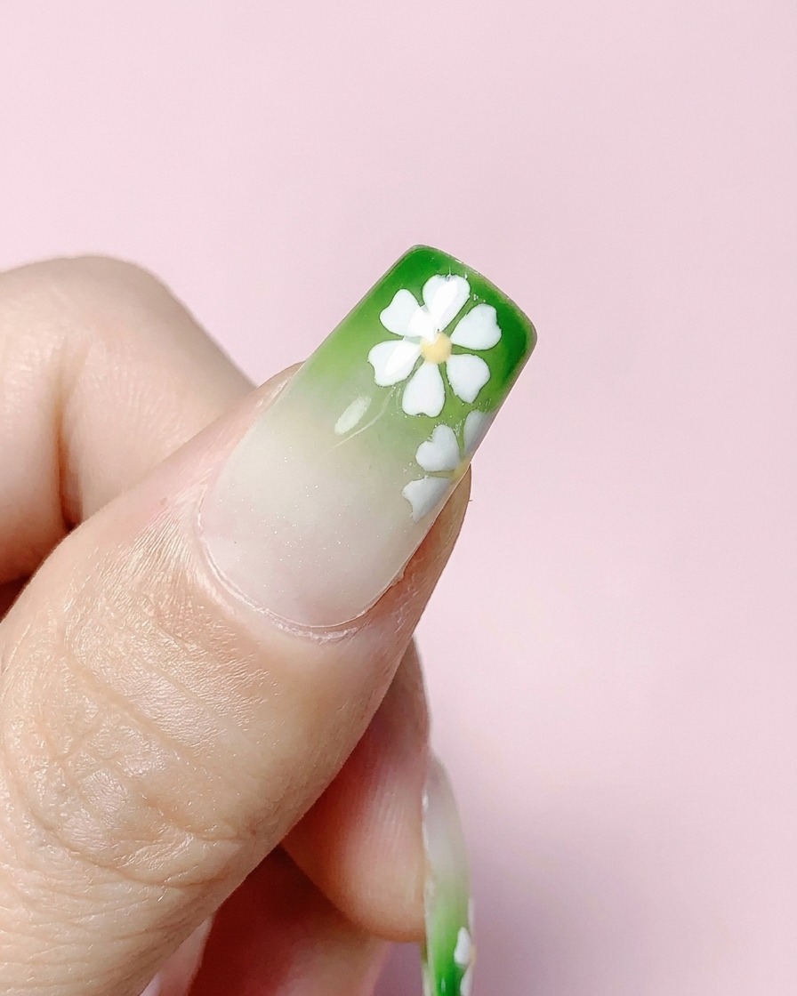 A finished square nail with green gradient and white flowers being coated with a clear top coat.