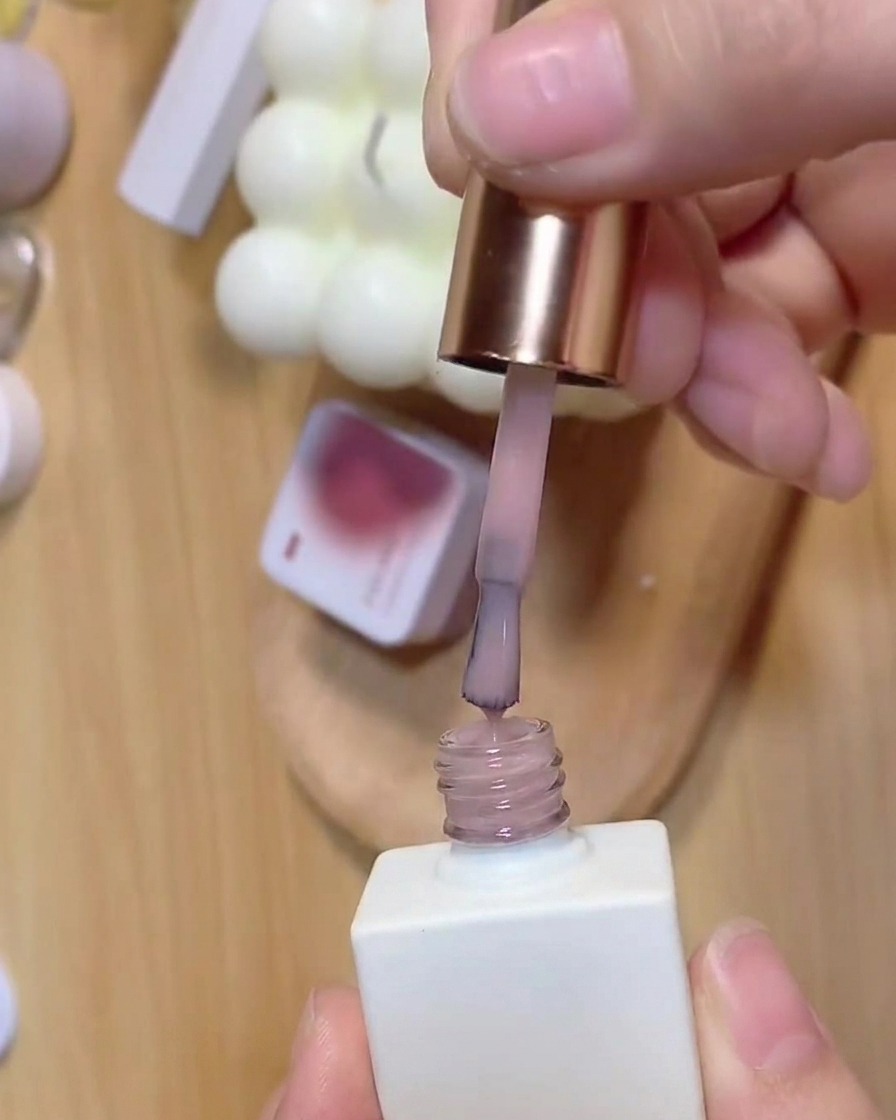 A brush pulling out translucent pink base coat from a white nail polish bottle.