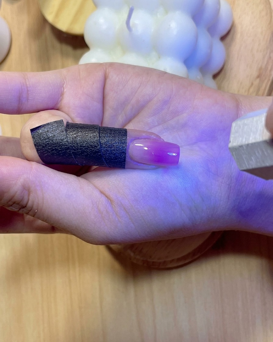 A fingernail glowing under purple UV/LED light while curing the applied reinforcement gel.