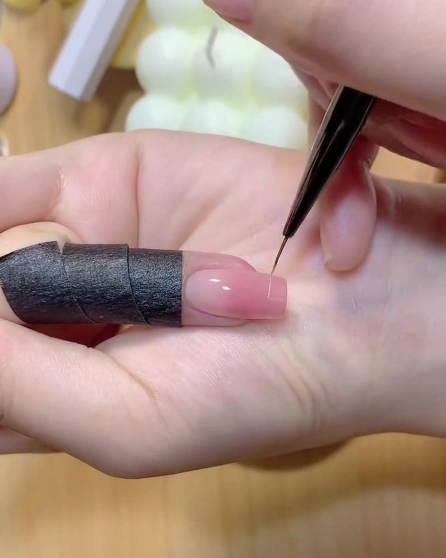 A fine liner brush drawing a thin white line across the tip of a pink gradient fingernail.