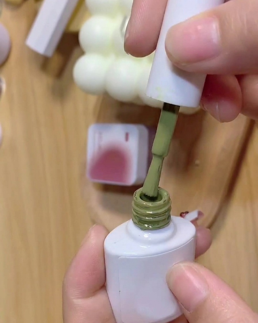 A brush dipping into a bottle of green gel polish with a background jar of pink gel.