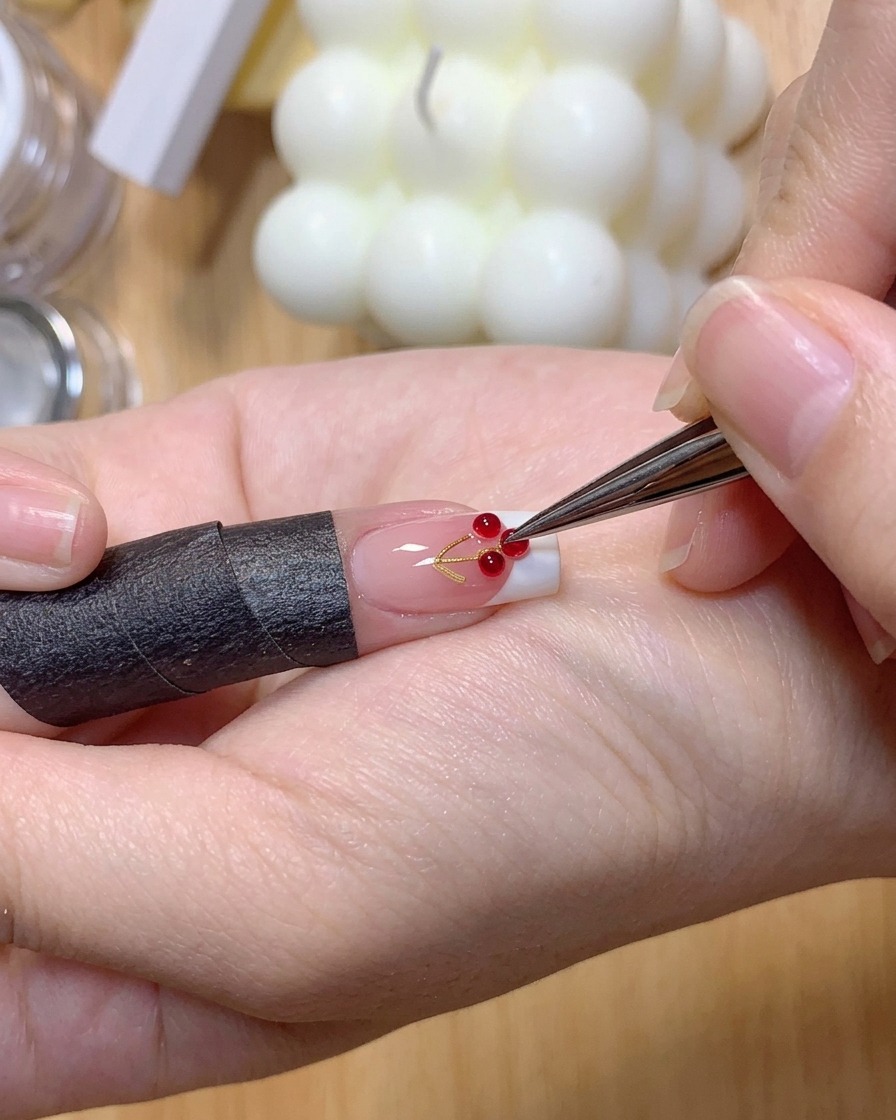 A pair of tweezers placing two small clear 3D gel balls onto a French manicured nail surface.