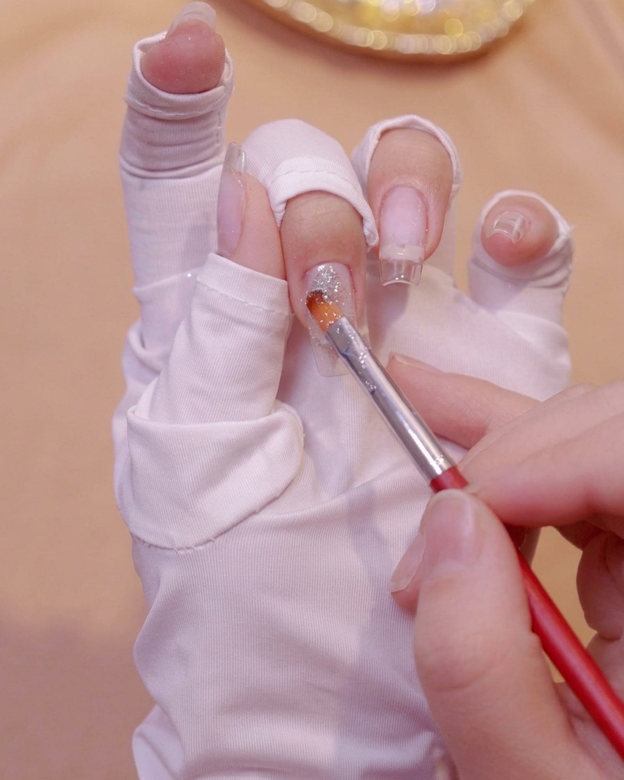A fine brush applying silver glitter gel near the base of a clear nail extension on a gloved hand.